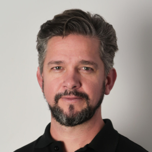 A portrait of a middle-aged man with short gray hair and a beard, wearing a black shirt, against a light gray background, sponsored by ATEC Australia International Ltd.
