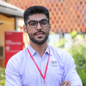 A man with glasses wearing a light blue shirt and a red GSMA lanyard stands outdoors with a modern building in the background.