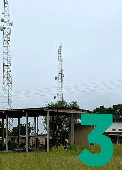 Two tall telecommunications masts with aerial dishes stand behind a partially constructed or open-sided building with a tin roof. There is green grass in the foreground, trees in the background, and a large green number 3 overlays the bottom right corner.