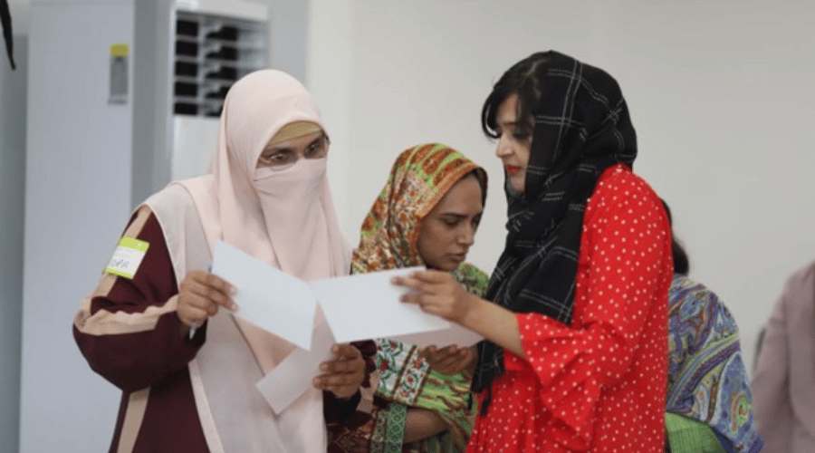 Three women, wearing colourful patterned clothes and headscarves, stand indoors. One woman in a niqab and glasses holds papers; another woman in a red polka-dot shirt with a black scarf looks at the papers; a third woman looks downward.