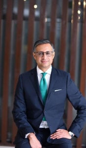 A man wearing glasses is seated, wearing a dark suit, white dress shirt, and teal tie. He has one hand on his knee and a slight smile. The background features vertical wooden slats and a blurred interior.