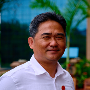 Man in a white shirt smiling outdoors with blurred greenery and buildings in the background.