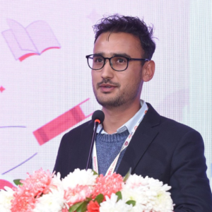 Man with glasses speaking at a conference, standing behind a podium with microphones and floral decorations, with a presentation screen in the background.