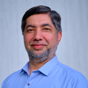Portrait of a middle-aged man with short dark hair, smiling, wearing a light blue shirt against a light gray background.