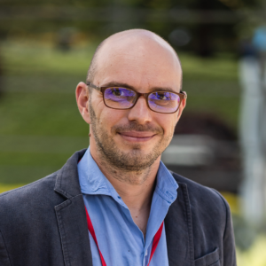 A man with a beard wearing glasses, a blue shirt, and a blazer smiles gently in an outdoor setting.