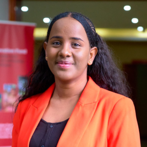 A professional woman in an orange blazer smiling at the camera, standing indoors with a blurred red background, representing the GSMA Innovation Fund for Anticipatory Humanitarian Action.