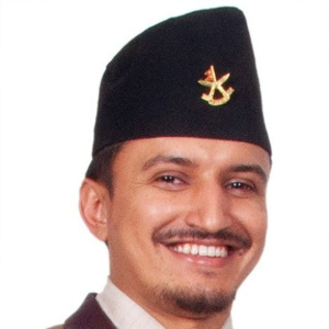 Man smiling, wearing a dark traditional cap with a gold emblem, dressed in a maroon outfit with a white collar.