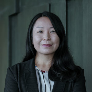 Aloi, with their long dark hair, stands poised against a dark, textured wall. Dressed in a pinstripe suit jacket and striped blouse, Aloi's expression is neutral, exuding a professional appearance that commands attention.