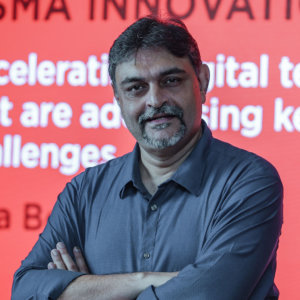 A person with short gray hair and a beard stands confidently with arms crossed in front of a red backdrop featuring white InQube text. They're dressed in a stylish gray button-up shirt.
