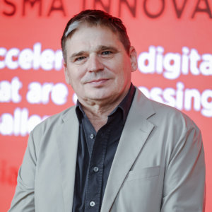 A man in a beige blazer and dark shirt stands confidently in front of a vibrant red backdrop, where partial text intriguingly hints at the Monsoon Tea Company's latest offerings.