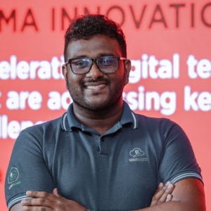 A person with glasses and a beard is wearing a dark polo shirt emblazoned with SenzAgro logos on the sleeves. They stand confidently, arms crossed, against a striking red background with partially visible white text.