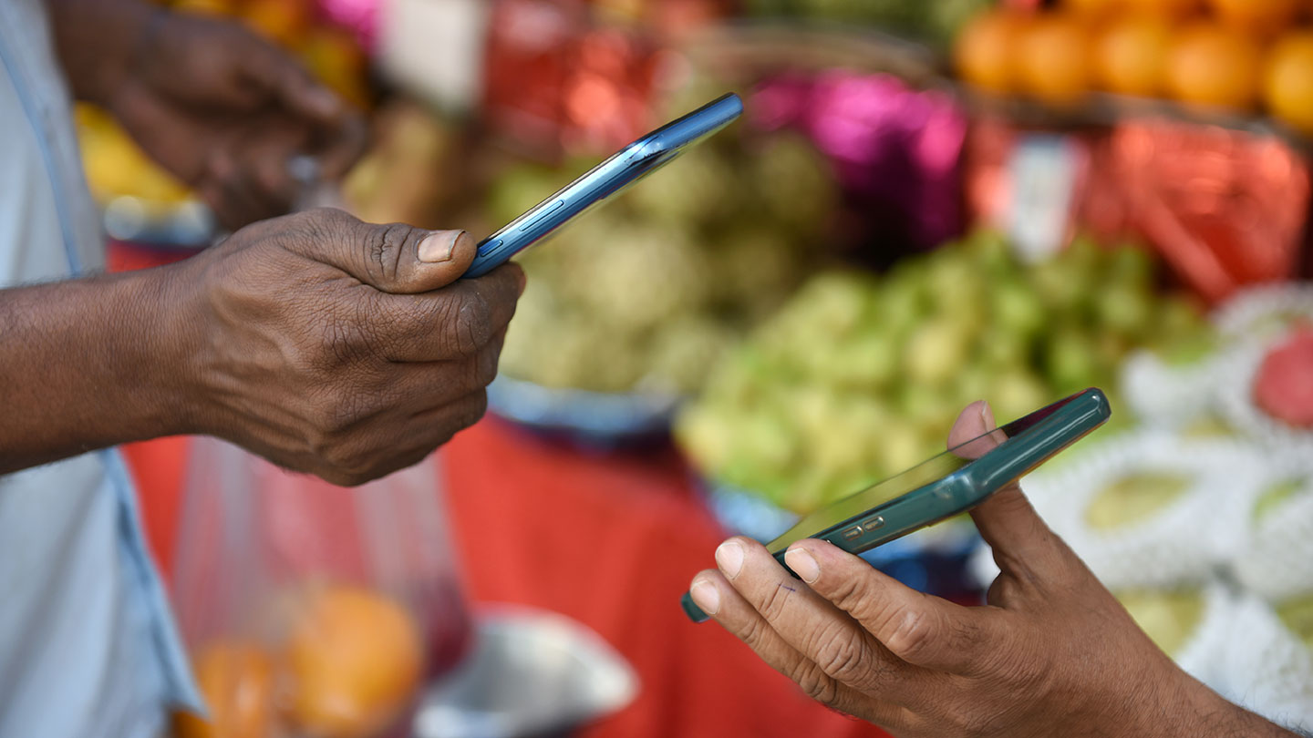 Two individuals using smartphones at a market with fruits in the background.
