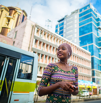 Woman waiting at a bus stop in an urban setting with modern buildings in the background.