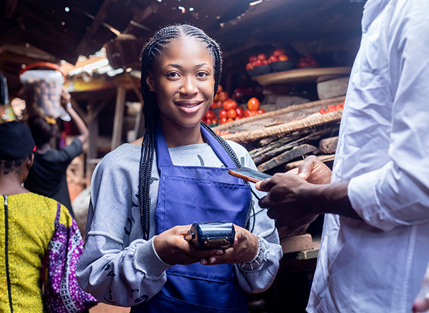 A smiling vendor in a blue apron holding a payment device while a customer hands over a card at a market stall.