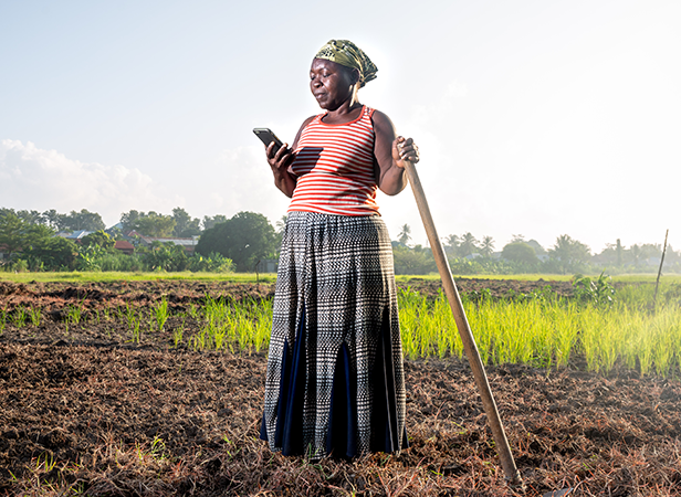 A farmer using a smartphone while standing in a field with a hoe in hand.