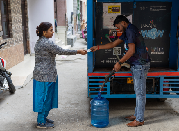 A woman paying a delivery man as he fills a large water jug from a mobile water distribution truck.