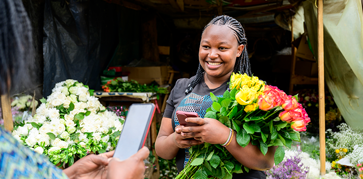 Customer using a smartphone to make a payment to a smiling florist, connected women, holding a bouquet of flowers.