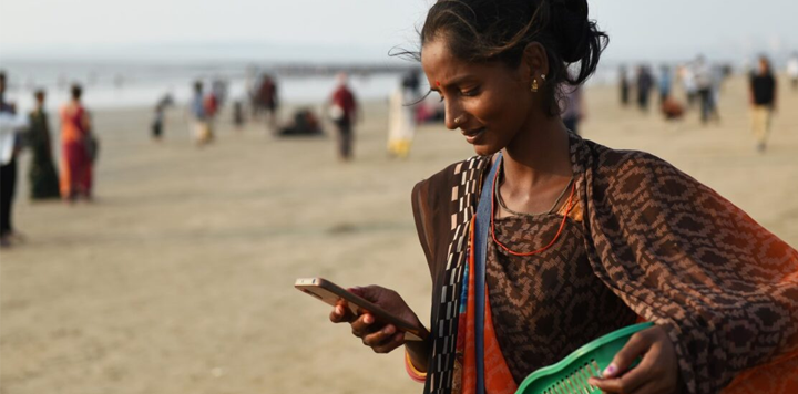 A woman looking at her mobile phone on a busy beach, experiencing the benefits of a GSMA Connected Society.