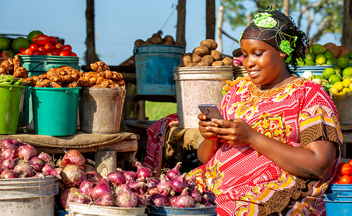 A woman using a smartphone at a colorful vegetable market stall.