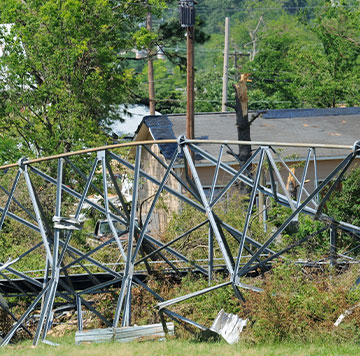 Collapsed metal tower with debris scattered on the ground amid trees and a building in the background.