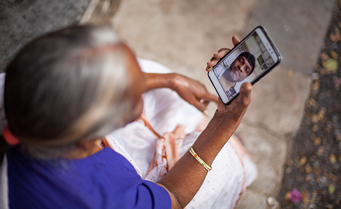 An elderly woman seated, looking at a mobile phone displaying a person's image on the screen.
