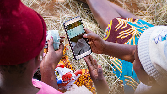 Two individuals sitting and looking at a smartphone together outdoors, engaged in a GSMA Mobile for Development initiative.