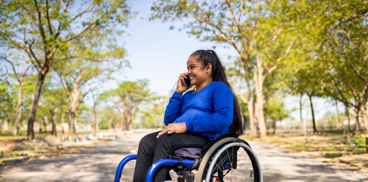 A woman in a wheelchair smiling while talking on the phone outdoors.