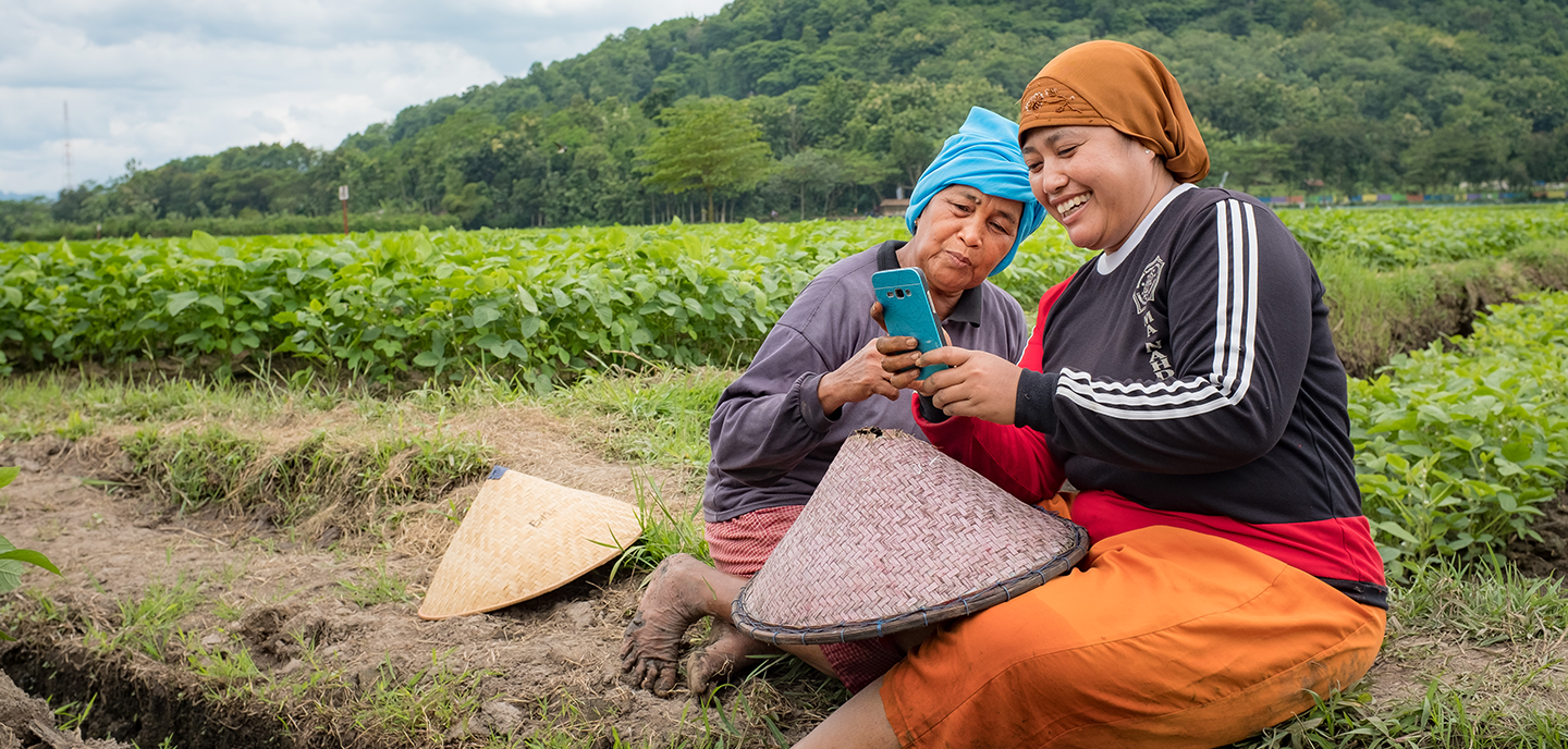 Two women smiling and looking at a mobile phone in a rural farm setting.