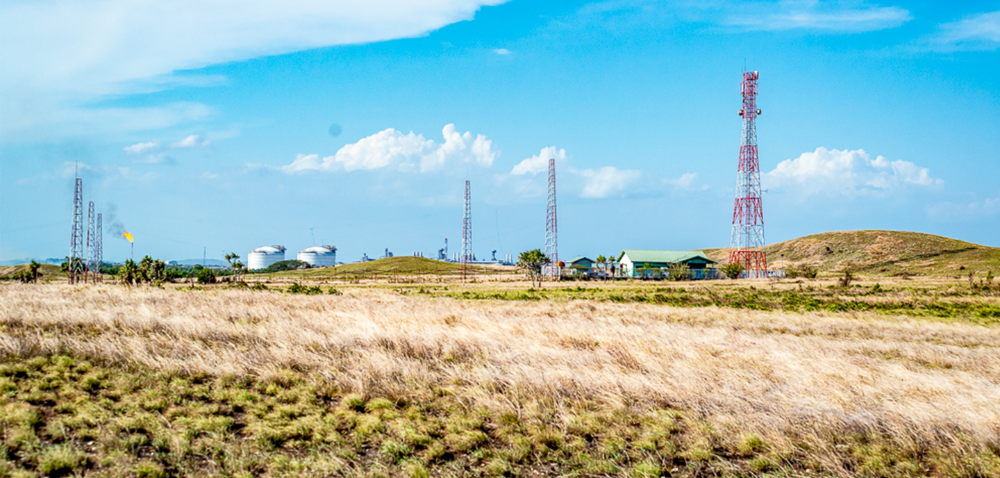 Rural industrial landscape with cellular towers, storage tanks, and sparse vegetation under a blue sky.