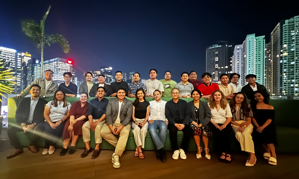 Group of people posing for a photo at a rooftop gathering during the evening with city buildings in the background.