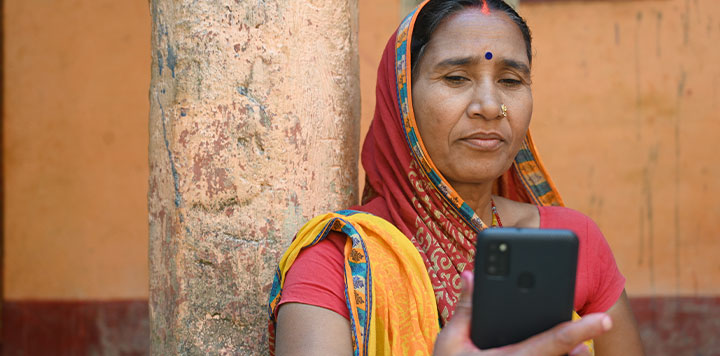 An Indian woman in a colourful sari uses a smartphone, leaning against a tree with a thoughtful expression.