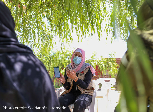 A woman wearing a mask and a hijab is sitting outdoors and presenting information on humanitarian innovation on a smartphone to another person whose back is to the camera, under the shade of green foliage.