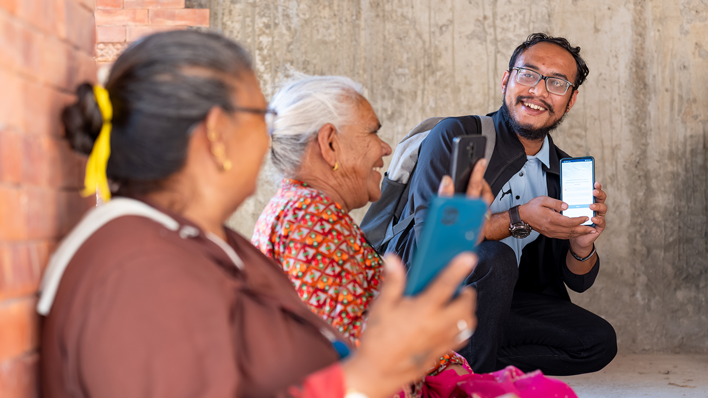 Young man showing smartphone screen to elderly women, all smiling during an informal gathering.