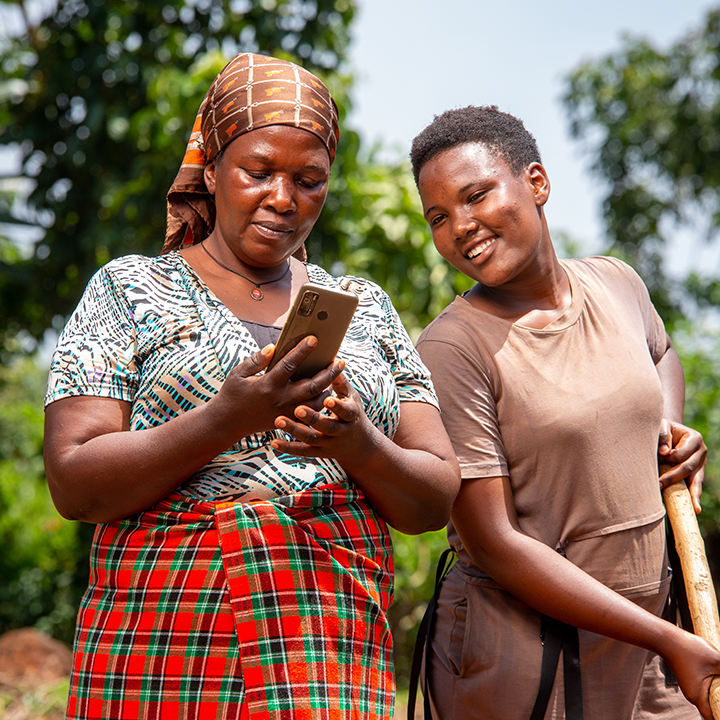 Two women in a rural setting, one holding a smartphone and the other with a hoe over her shoulder, looking at the phone screen together.