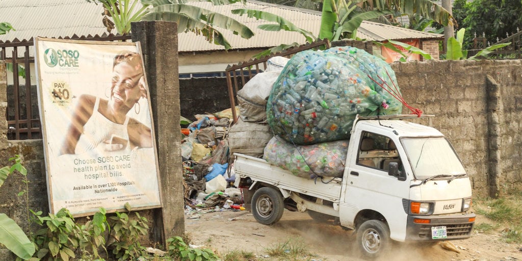 A small white truck carrying large, bulging bags of plastic bottles drives through a dusty pathway next to a brick wall. A health insurance advertisement featuring a smiling woman is displayed on the wall to the left.