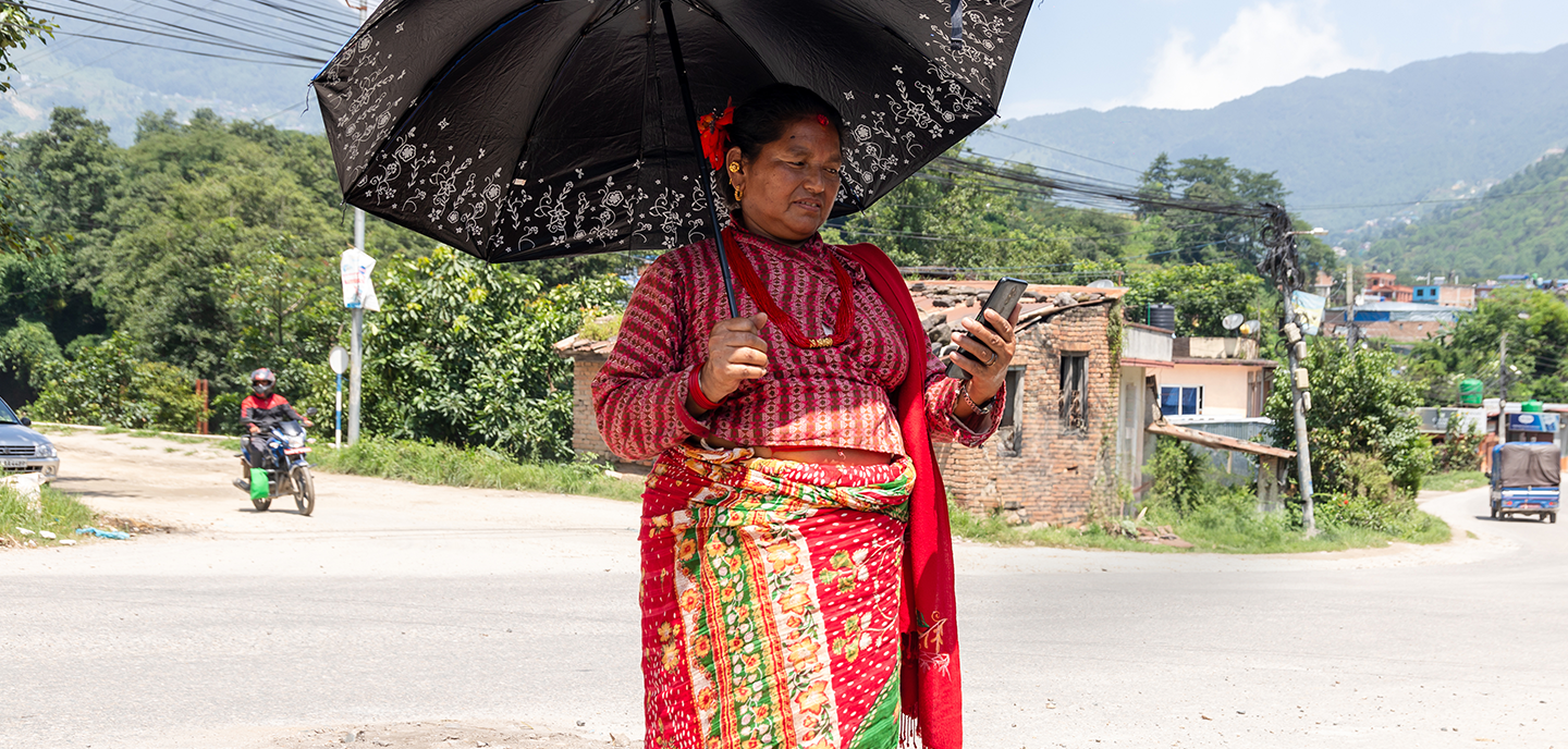 A woman stands on a roadside, holding a black umbrella while looking at her smartphone, exemplifying the connected women of today. Dressed in traditional colorful clothing with a red and green saree, she stands against a backdrop of motorbikes and buildings.