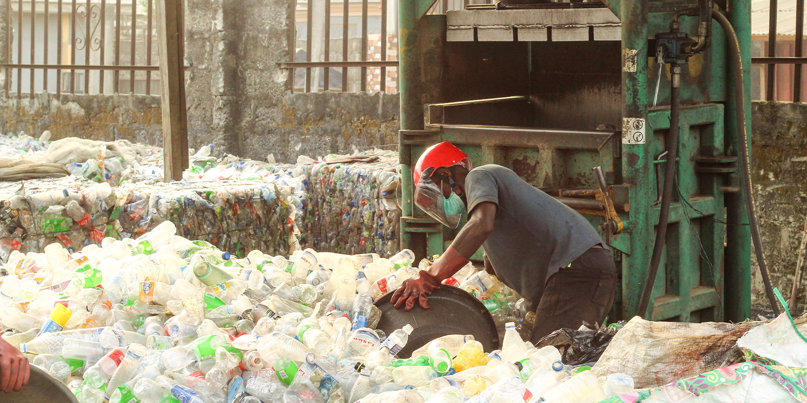 A person wearing a red helmet and a face mask is seen pushing recyclable plastic bottles into a green machine at a recycling facility. Large quantities of plastic bottles are scattered on the ground around them.
