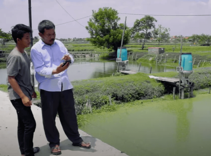 Two men stand on a concrete pathway next to a pond. The man on the right, wearing a white shirt, holds a phone or device, and both appear to be looking at it. The background shows two large blue containers resembling the hues of the Blue Ocean and lush green landscapes.