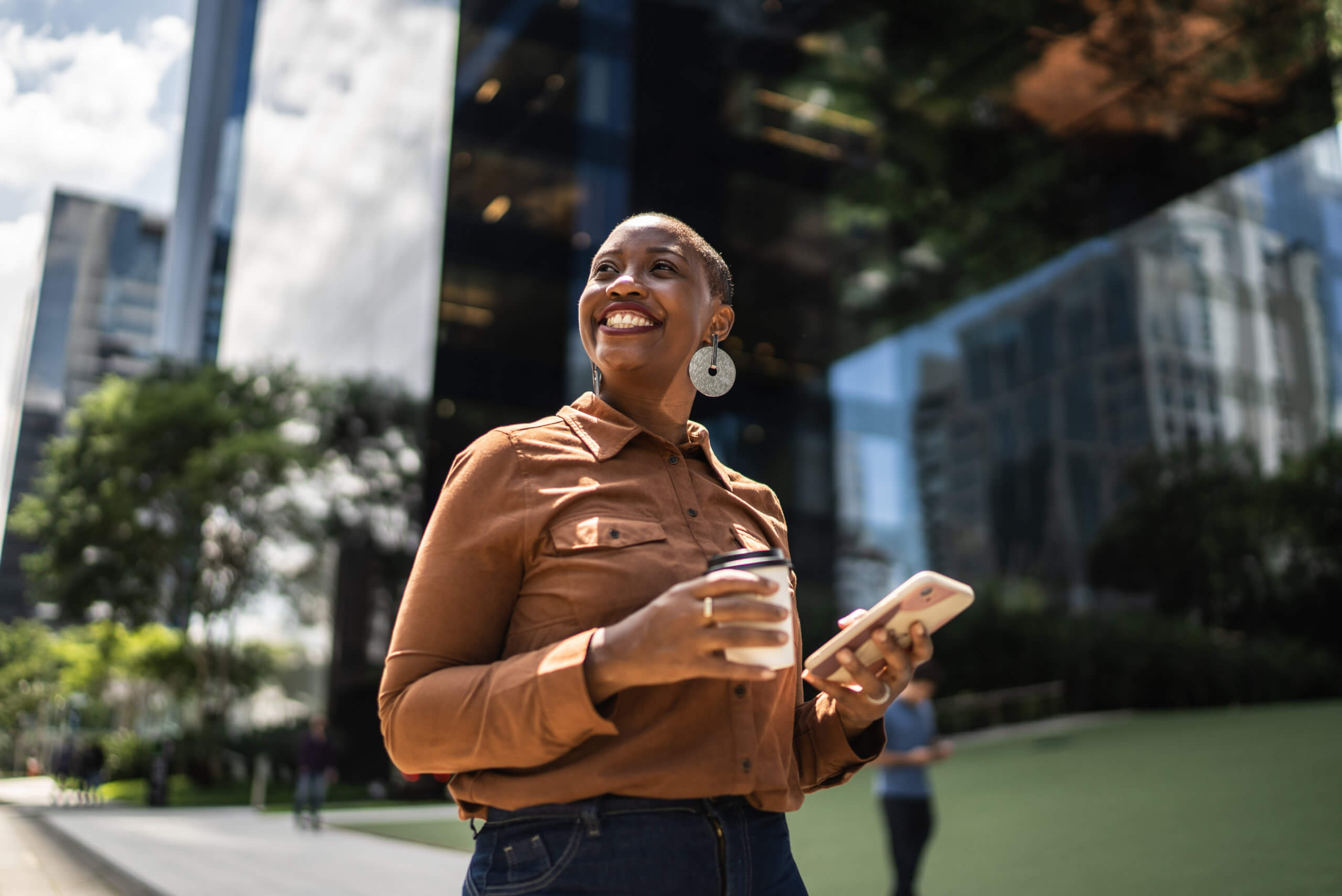 A person is standing outside in an urban area, holding a coffee cup in one hand and a smartphone in the other. They are smiling and looking off into the distance. Behind them are tall, reflective buildings and some greenery. The weather appears to be sunny.