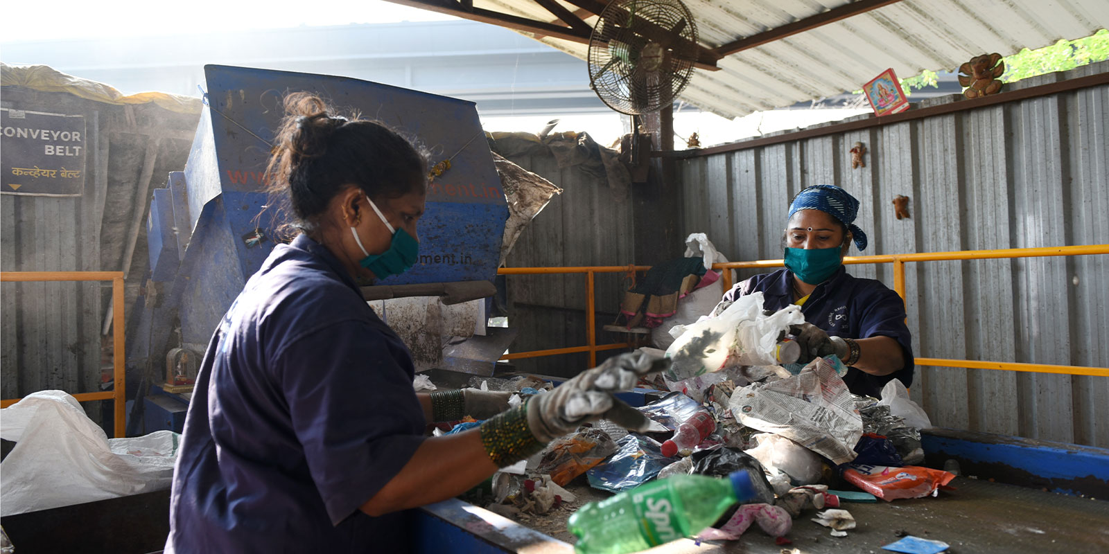 Two women wearing blue uniforms and face masks are sorting through garbage on a conveyor belt under a covered structure. One woman holds a green plastic bottle. Various types of waste are scattered on the belt. The background includes metal walls and equipment.
