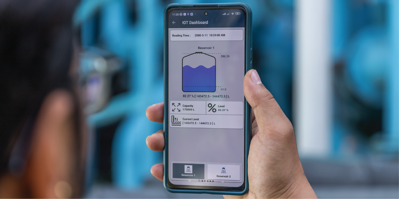 A person is holding a smartphone displaying an IoT Dashboard for a reservoir. The screen shows the reservoir is 82.7% full, with current water level, capacity, and usage details. In the background, there are blurred blue-colored industrial components.