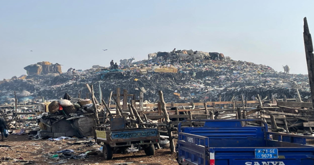 A large pile of garbage in a landfill with people and animals scavenging on top. Various wooden carts and a blue motorized cart are in the foreground near the waste. The sky is hazy, and the scene appears chaotic and cluttered.