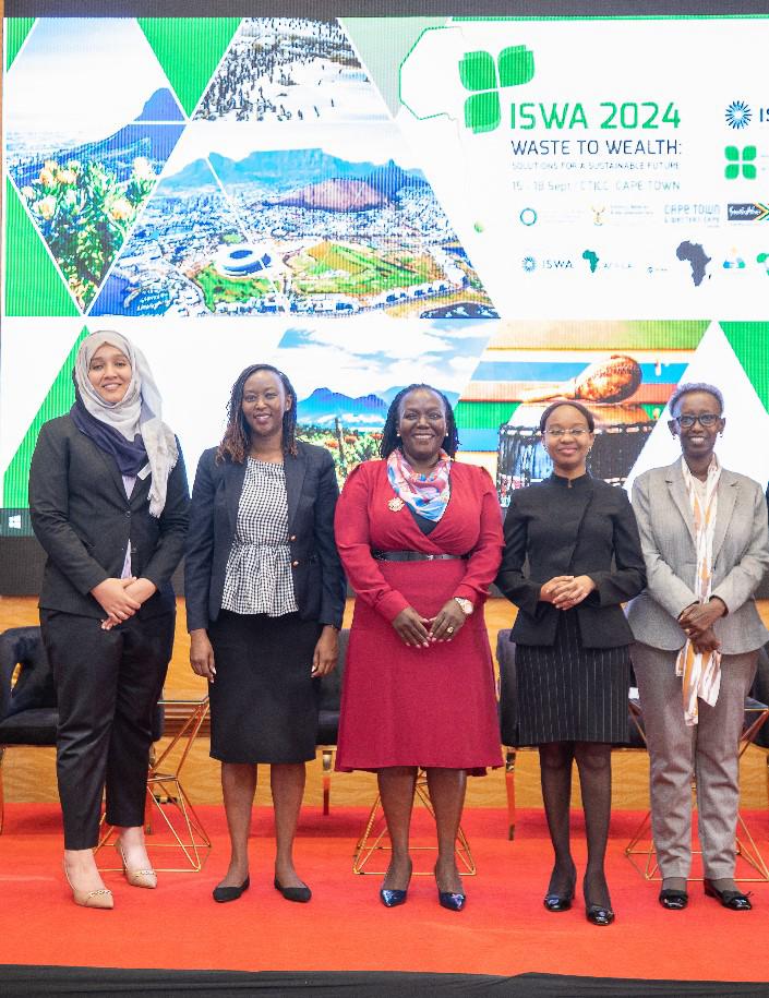 Five women are standing in a row, smiling at the camera in front of a projected screen displaying information about the ISWA 2024 conference with the theme "Waste to Wealth." The background includes images of landscapes and event details.