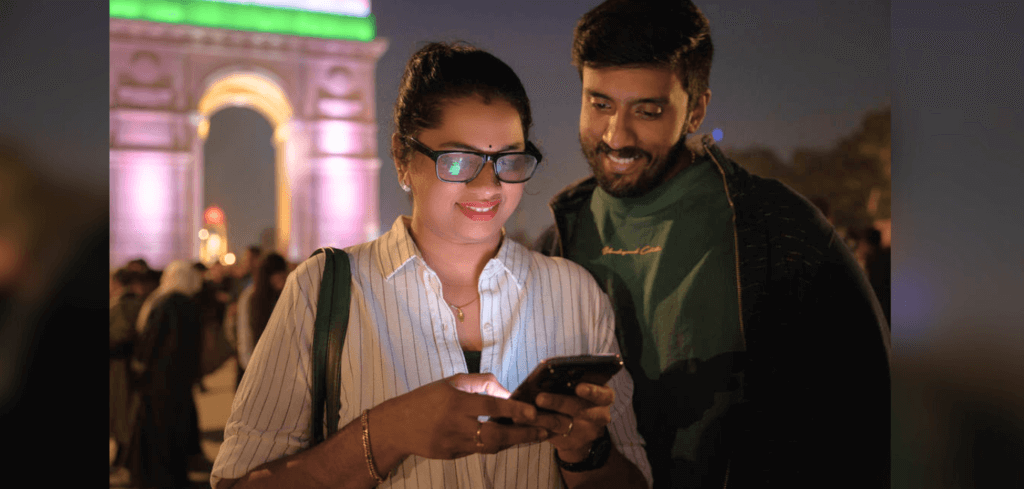 Two people are standing outdoors at night near a brightly lit archway. The person on the left is holding and looking at a smartphone, smiling, while the person on the right is looking at the same phone and smiling. They are surrounded by other people.