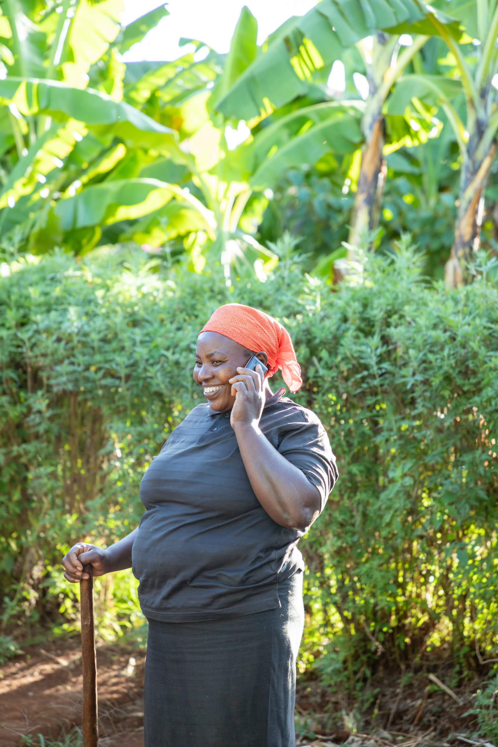 A woman stands in her field, holding a farming tool in one hand and her mobile phone in the other. Using mobile money and her digital financial literacy skills, she accesses credit to invest in better equipment and grow her agricultural business.