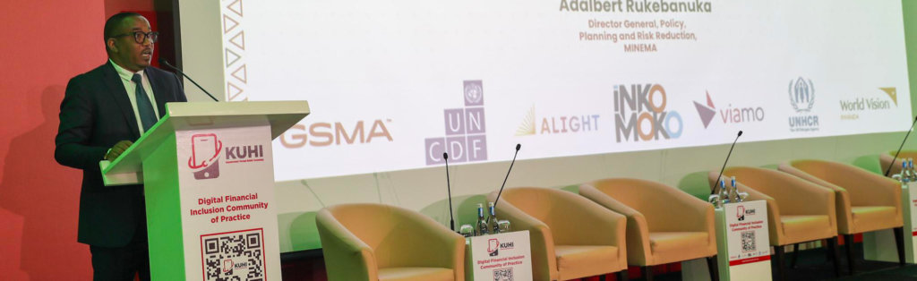 A man in a suit speaks at a podium on a stage with a large screen behind him displaying various organization logos and text. Four beige chairs with microphones are arranged on stage, each chair with a name placard and a QR code stand in front.