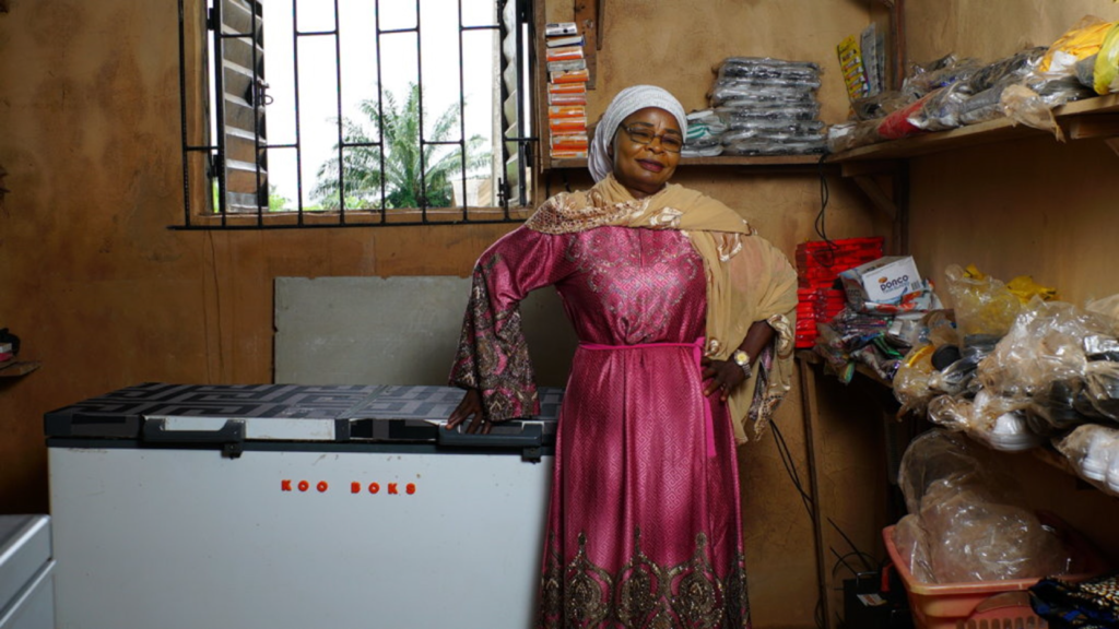 A woman in a pink and gold dress with matching headscarf stands confidently beside a large freezer labeled "Koo Boks" in a small, well-stocked shop. Shelves hold various packaged goods. A window with bars reveals greenery outside.