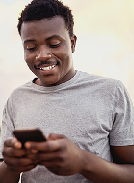 A person wearing a gray T-shirt is smiling while looking at a smartphone. The background is softly blurred, highlighting the person's expression and focus on the device.