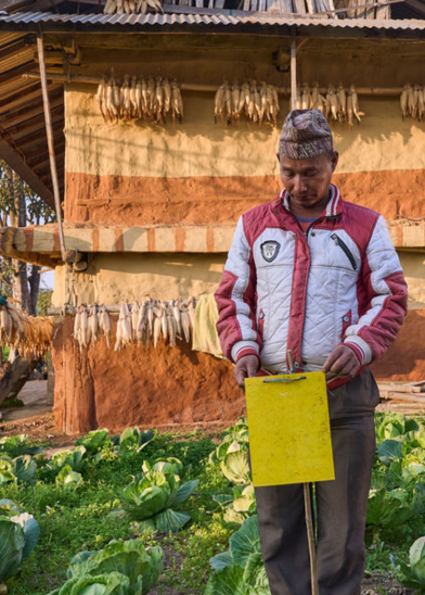A man in a red and white jacket stands in a garden with cabbage plants, holding a yellow board with "GSMA Green Glossary" written on it. Behind him is a rustic building adorned with corncobs hanging from the eaves, reflecting a commitment to eco-friendly practices.