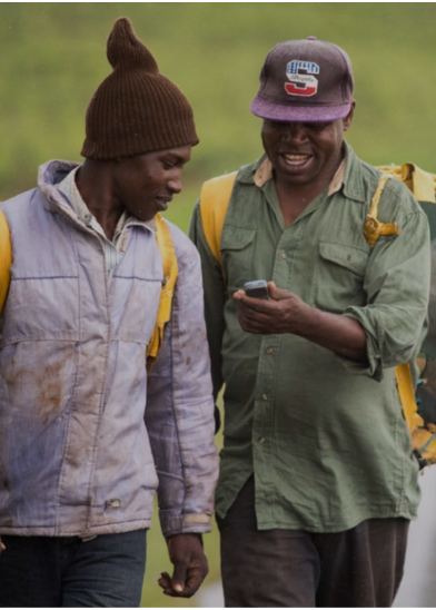 Two men stroll along a grassy path, backpacks in tow. One, clad in a green shirt and baseball cap, smiles as he shows something from the GSMA Green Glossary on his phone to his friend in a brown beanie and light jacket.